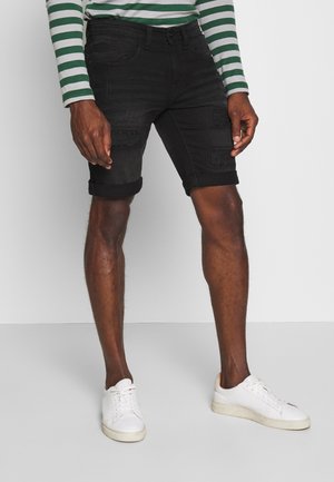 Zwarte denimhorts met verwerkte details en opgerolde manchetten, gecombineerd met witte sneakers. Model draagt een groen-witte gestreepte lange mouwen shirt.