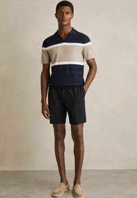 Polo shirt tricotat cu dungi bleu marin și bej, cu mâneci scurte, asortat cu pantaloni scurți negri și pantofi slip-on bej, expus pe un fundal neutru.