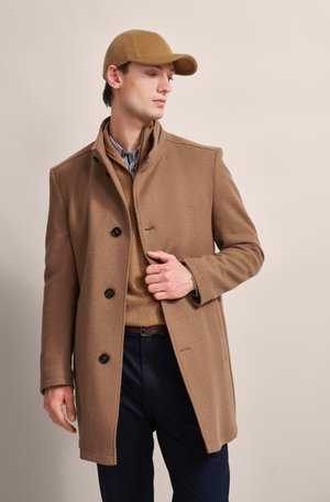 Brown wool blend coat with a high collar, button closure, and side pockets, layered over a beige knit sweater and blue trousers.