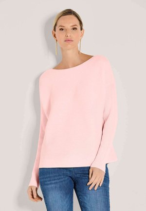 Vrouw met een lichte huid die een lichtroze top met lange mouwen en blauwe spijkerbroek draagt, staand tegen een effen beige achtergrond.