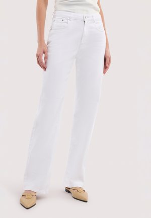 Frau trägt weiße, hoch taillierte, gerade geschnittene Jeans und beige Mules mit spitzem Zehenbereich und goldenen Schnallendetails, steht vor hellem Hintergrund.