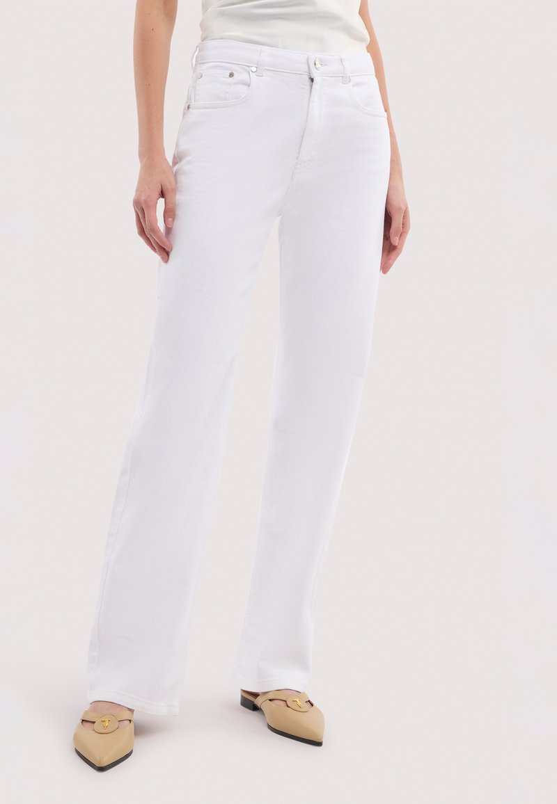 Femme portant un jean blanc taille haute à jambes droites et des mules beiges à bout pointu avec des détails de boucle dorée, debout sur un fond clair.