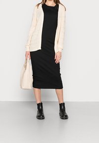 Krämbelädd cardigan i kombination med en svart ribbad midiklänning, svarta ankelkängor och en ljusbeige handväska, på en grå golv bakgrund.