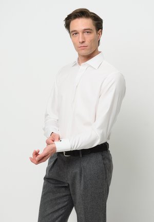 Witte lange mouwen overhemd met een puntkraag en knoopmanchetten, gecombineerd met grijze op maat gemaakte broek. Het model past de manchet aan.