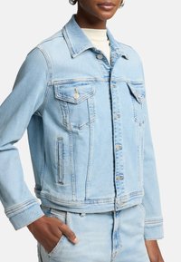 Ljusblå denimjacka med knäppning framtill, två bröstfickor och sidofickor; har en klassisk krage och sydda detaljer på ärmarna.