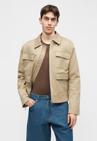 Νεαρός άνδρας που φορά μπεζ ζακέτα με φερμουάρ και μεγάλες τσέπες με καπάκι πάνω από καφέ πουκάμισο και μπλε τζιν, στέκεται με το ένα χέρι στην τσέπη.