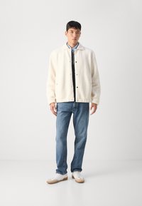 Fløjl-lined cremet jakke med trykknapper, parret med blå jeans og hvide sneakers. Jakken har krave og et stilrent design.