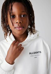 Fehér kapucnis pulóver fekete "ALLSAINTS" logóval a zsebnél, húzózsinóros kapucnival és bordázott mandzsettákkal. A modellen barna bőr és raszta frizura látható.