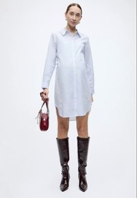 Hellblau gestreiftes Hemdblusenkleid mit langen Ärmeln, durchgehender Knopfleiste, seitlichen Schlitzen, kombiniert mit glänzenden schwarzen kniehohen Stiefeln und einer kleinen roten Tasche.