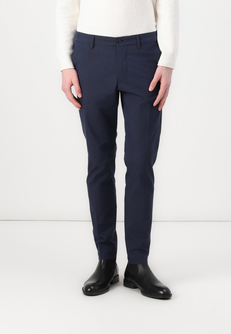 Sötétkék, könnyű anyagból készült nadrág, slim-fit szabású, elöl gombzáras és oldalsó zsebekkel. Fekete bokacsizmával párosítva.