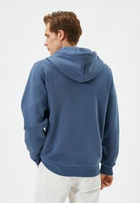 Hombre con cabello castaño claro vistiendo una sudadera azul y pantalones blancos, de pie de espaldas con las manos en los bolsillos, contra un fondo blanco liso.