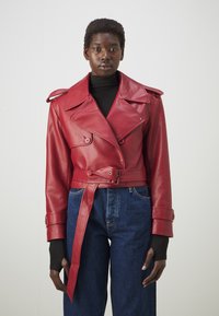 Rød læder trenchcoat med store revers, dobbeltknaplukning, bæltedetalje i taljen og skulderepauleter, båret over mørke denimbukser.