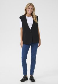 Črn brezrokavnik z izrezanim ovratnikom, enojnim gumbom, dvema stranskima žepoma, oblečen preko belega majice in modrih jeans hlač.