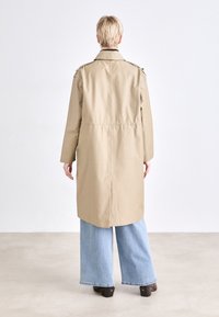 Μπεζ παλτό με χαλαρή εφαρμογή, που διαθέτει επιθέματα ώμου, σφικτή μέση και άνοιγμα στην πλάτη. Φοράει πάνω από ανοιχτό μπλε τζιν σε φαρδιά περικοπή.