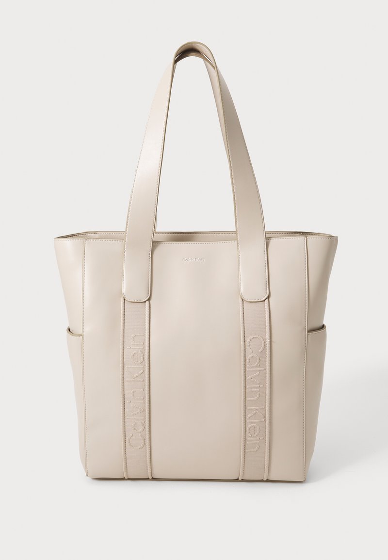 Beżowa torebka typu tote z sztucznej skóry z podwójnymi płaskimi uchwytami i wytłoczonym logo z przodu. Posiada pionowe przeszycia oraz otwarte kieszenie po bokach.