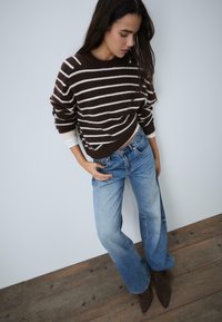 Brązowy sweter w paski o luźnym kroju, zestawiony z jasnoniebieskimi spodniami z szerokimi nogawkami oraz ciemnobrązowymi botkami za kostkę, na neutralnym tle.