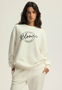Sweter w kolorze kremowym z czarną okrągłą grafiką z napisem "Bloovii" oraz dodatkowym tekstem poniżej. Luźny krój i miękka tkanina.