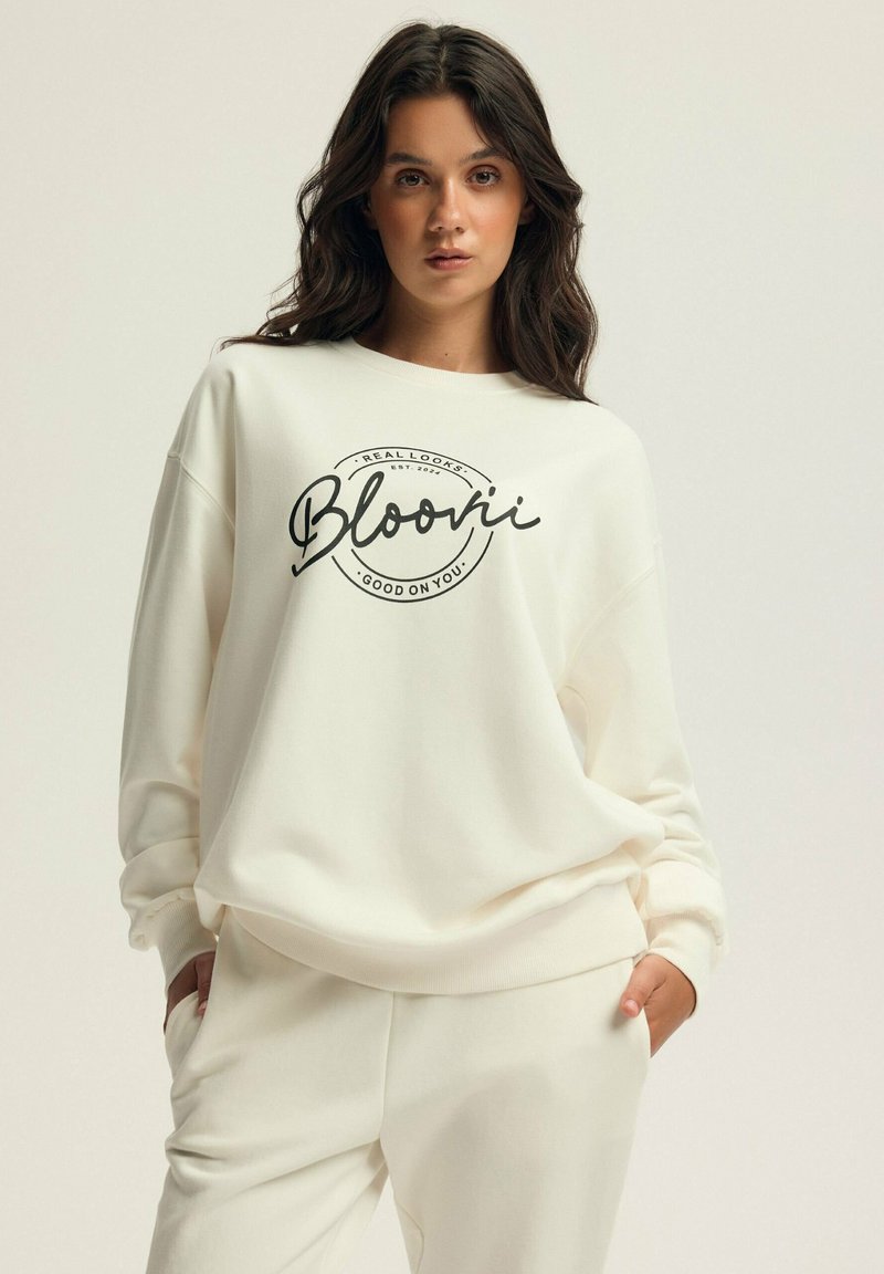 Sweter w kolorze kremowym z czarną okrągłą grafiką z napisem "Bloovii" oraz dodatkowym tekstem poniżej. Luźny krój i miękka tkanina.