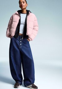 Donna che indossa un giubbotto imbottito rosa chiaro sopra un hoodie nero, una maglietta bianca a maniche corte, jeans larghi blu e sneaker scure, in piedi con le mani nelle tasche.