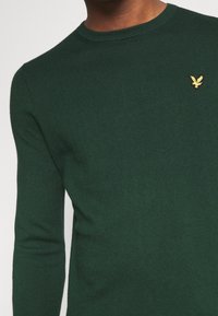 Pull en maille à manches longues de couleur vert foncé avec un petit emblème d'oiseau jaune sur la poitrine gauche, porté par une personne à la peau de ton moyen.