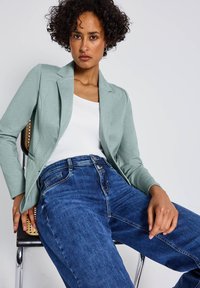 Blazer vert clair sur un top blanc, associé à un jean en denim bleu. Le blazer présente un design épuré, et le jean a une coupe classique.