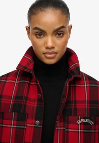 Red and black plaid flannel jacket with a fold-down collar, featuring two front pockets and a stitched logo on the left chest.