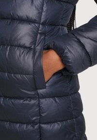 Marinblå puffjacka med quiltad isolering, en slät yta och en sidoficka med en synlig hand i för att visa skalan.