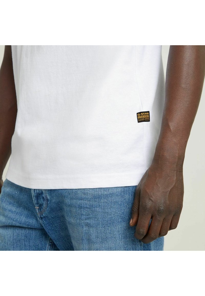 Camiseta de algodón blanca con escote redondo y mangas cortas. Presenta una etiqueta negra y amarilla de G-Star en el dobladillo. Llevada con pantalones vaqueros azules.