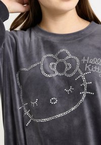 Grå langærmet skjorte lavet af blødt stof, med et Hello Kitty-design prydet med sølv rhinsten for detaljer.