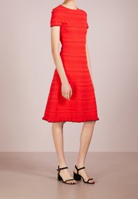 Femme portant une robe rouge texturée longueur genou avec des manches courtes et des sandales à talons blocs noires à lanières, se tenant sur un sol lisse.