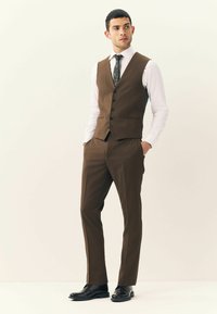 Costum maro, realizat pe măsură, cu o vestă ajustată cu nasturi, completat cu pantaloni asortați și pantofi eleganți negri, pe un fundal neutru.