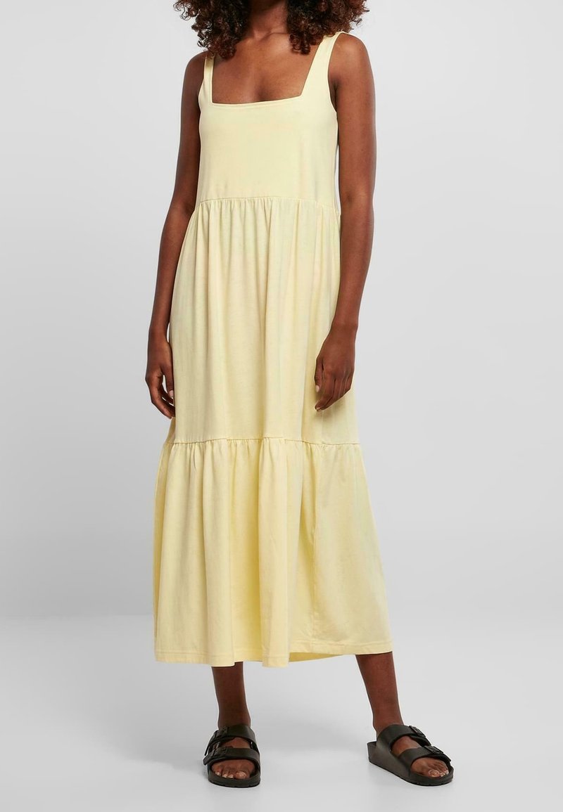 Femme portant une robe midi sans manches jaune pâle à étages et des sandales slides noires, debout devant un fond clair uni.