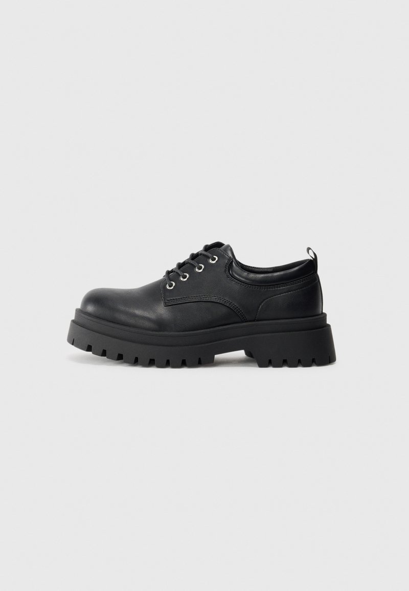 Zapatos de cuero negro con cordones, que presentan una suela de goma gruesa con un profundo taqueado, punta redondeada y ojales de metal. Textura suave en toda su superficie.