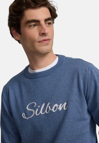 Suéter azul de material de punto suave con escote redondo y la palabra "Silbon" en script plateado en el pecho.