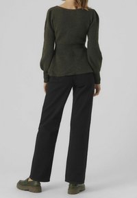 Donna che indossa un pullover in maglia verde scuro, pantaloni neri a gamba larga e scarpe platform verdi, in piedi di spalle.