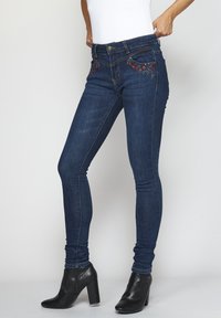 Kapeat farkut tummassa denimissä, joissa on koristeellinen kukka-ompelu taskuissa. Ominaisuuksiin kuuluu keskihäntä ja standardi viisi-taskuinen muotoilu. Mustat koroilla varustetut nilkkasaappaat.