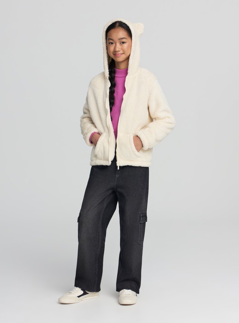 Kremowa polarowa kurtka na zamek z kapturem z uszami misia, zestawiona z fioletową bluzką z długim rękawem i czarnymi spodniami typu cargo.