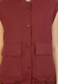 Burgundy ärmlös väst med v-ringning, framknappar och två stora bröstfickor. Texturerad tyg med en samlad fållsdetalj.
