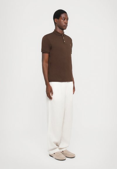 Camisa polo marrón de manga corta, con tapeta de tres botones; combinada con pantalones blancos holgados y zapatos color beige claro. Diseño simple y clásico.
