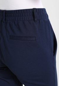 Námořnické modré kalhoty s elastickým pasem, obohacené o boční kapsu a hladkou texturu látky. Design zahrnuje jemné detaily prošívání.