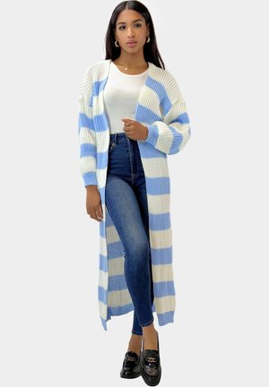 Frau trägt eine lange blau-weiß gestreifte Strickjacke, weißes Ripp-T-Shirt, dunkle Skinny-Jeans und schwarze Loafers, posiert vor einem einfarbigen Hintergrund.