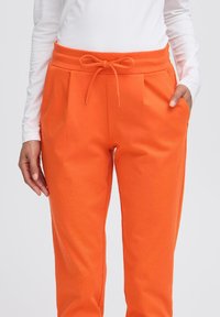 Oranje trainingsbroek met een elastische tailleband, plooien, zijvakken en een gladde, zachte textuur. Gecombineerd met een wit longsleeve shirt.