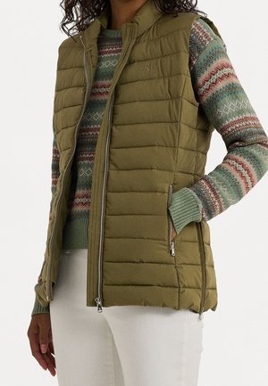 Veste matelassée vert olive avec fermeture éclair sur un pull en maille à motifs multicolores et un pantalon blanc, main dans la poche de la veste.