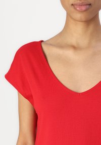 En ung person som bär en röd V-ringad T-shirt, som visar nyckelbenet och en del av nacken och läpparna, mot en enfärgad vit bakgrund.