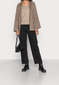 Blazer a quadri marroni sopra un top in maglia beige, abbinato a pantaloni larghi neri e stivaletti neri, mentre porta una borsa a spalla nera.