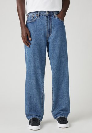 Personne portant un jean bleu ample, une chemise blanche, des baskets noires et blanches, debout avec une main dans la poche sur un fond gris.