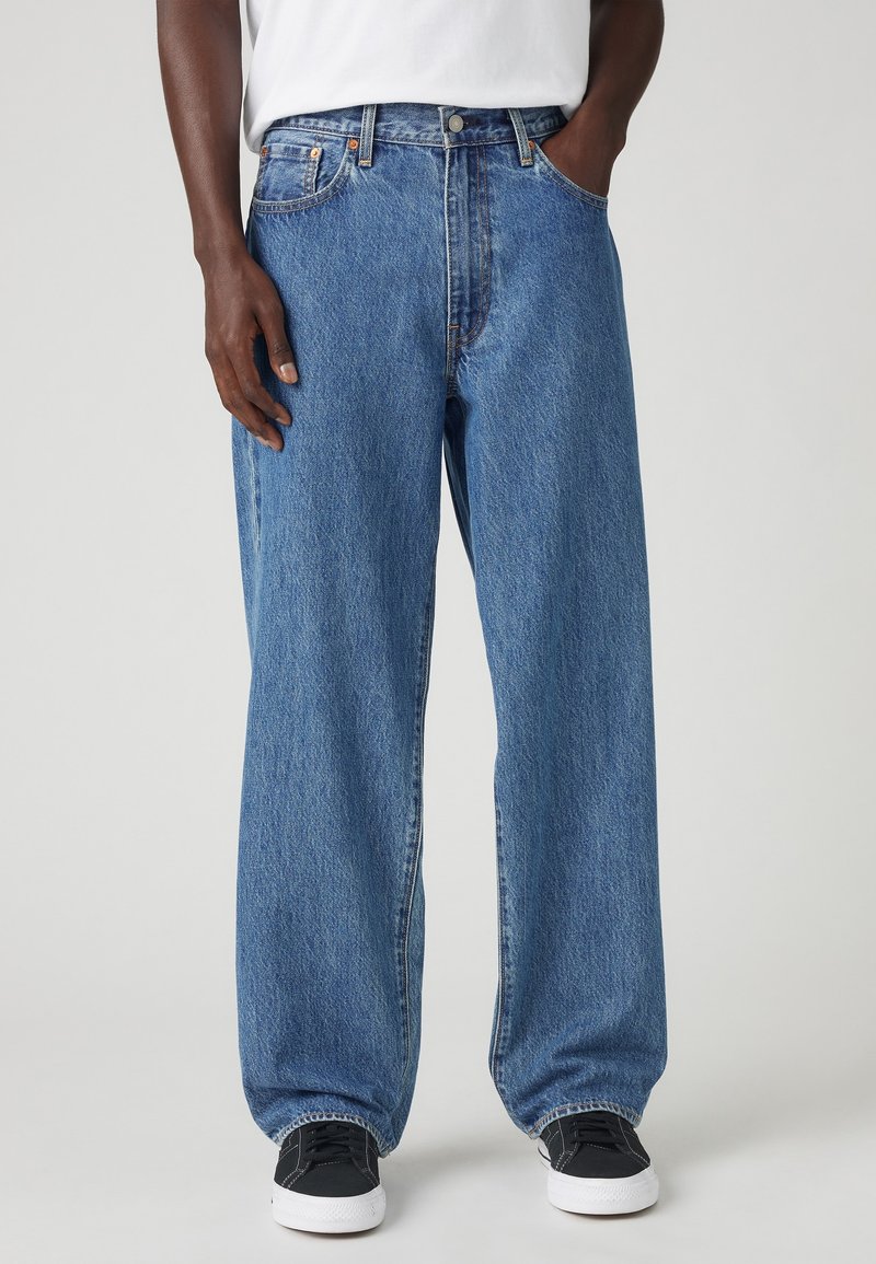 Personne portant un jean bleu ample, une chemise blanche, des baskets noires et blanches, debout avec une main dans la poche sur un fond gris.