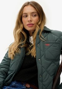 Chaqueta verde acolchada con un bolsillo y un logo, llevada sobre un suéter negro de punto. El modelo tiene el pelo rubio, con un maquillaje mínimo.