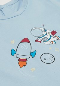 Sv�ětle modrá bavlněná košile s motivem rakety s červenými akcenty a návrhem astronauta, obklopená hvězdami a planetami. Jednoduchá, hravá grafika.