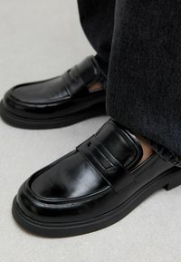 Mocassini neri in pelle con finitura liscia, dotati di una fascia sopra il collo del piede e una suola in gomma resistente, abbinati a pantaloni in denim scuro.